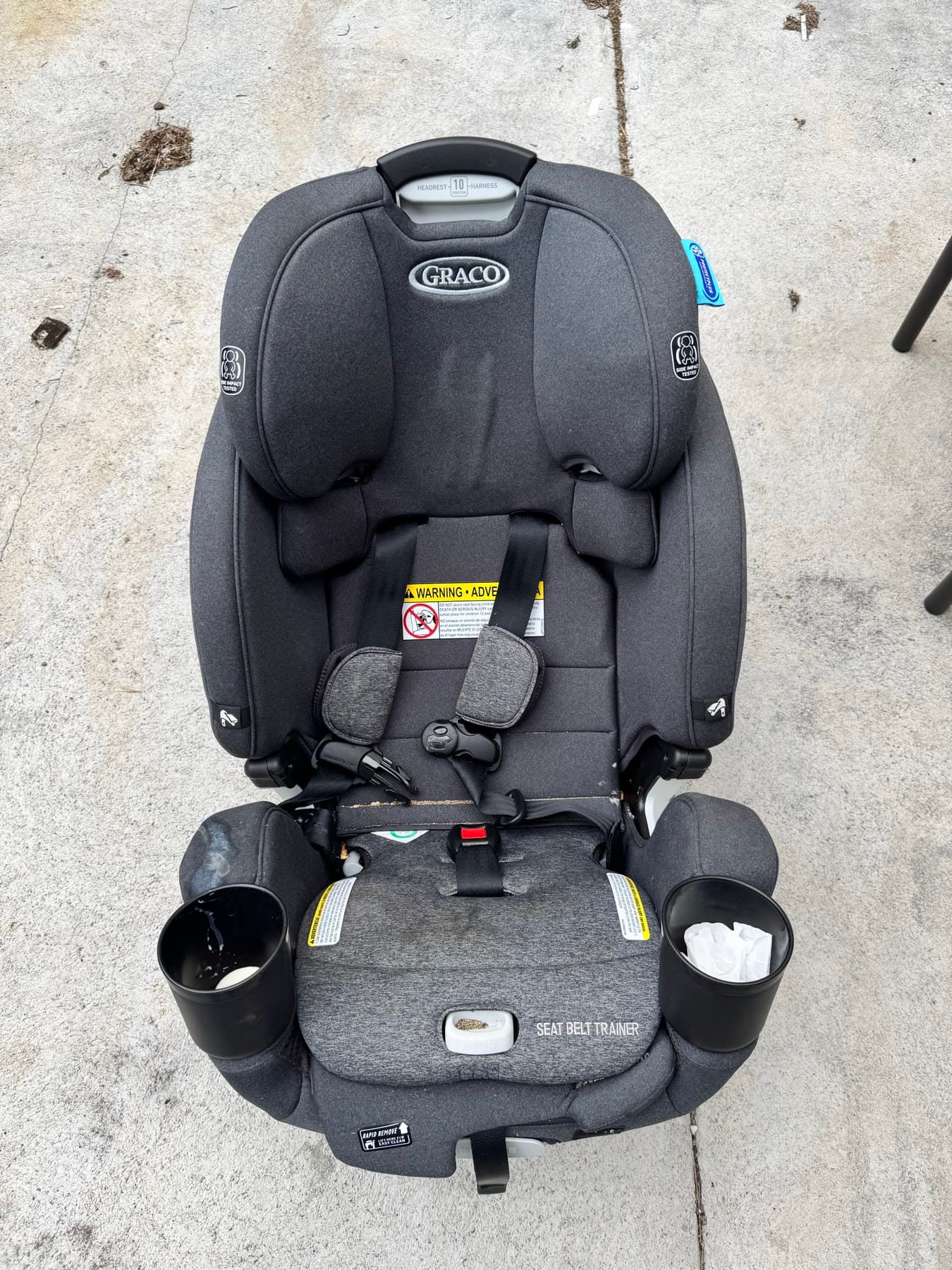 A grey Graco car seat photographed from above before cleaning, with dried milk in one cup holder and crumbs on the seat pad